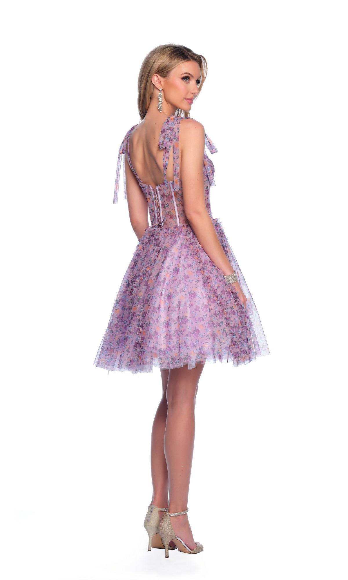 A woman poses against a white background, looking over her shoulder in the Dave and Johnny Short Print Hoco Dress 11752—a light purple, floral party dress with corset-style bodice, bow straps, open back—and nude high heels.
